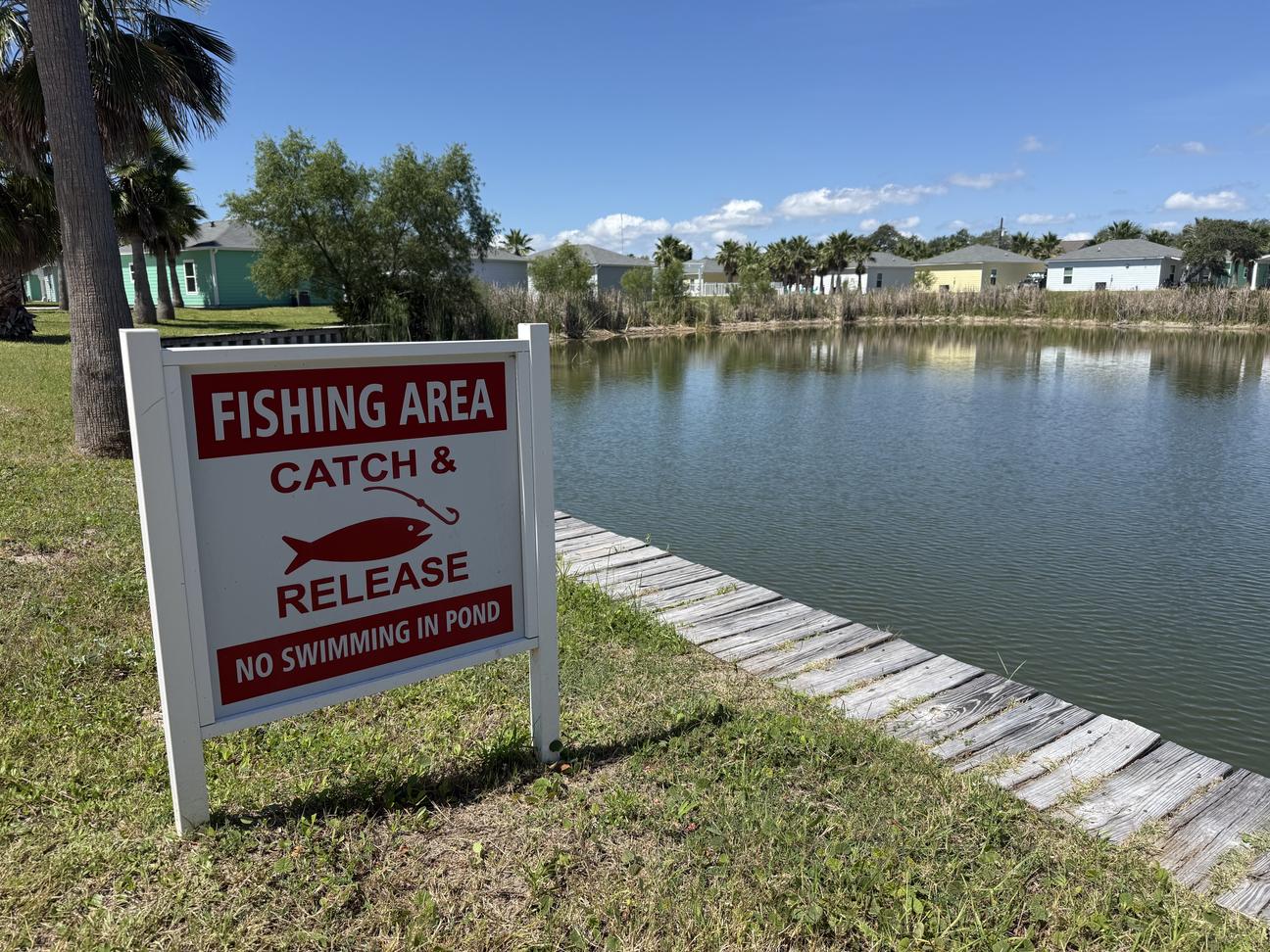 Catch-and-release fishing pond surrounded by coastal cottages, palm trees, and grassy shoreline with posted fishing area sign under a sunny blue sky.