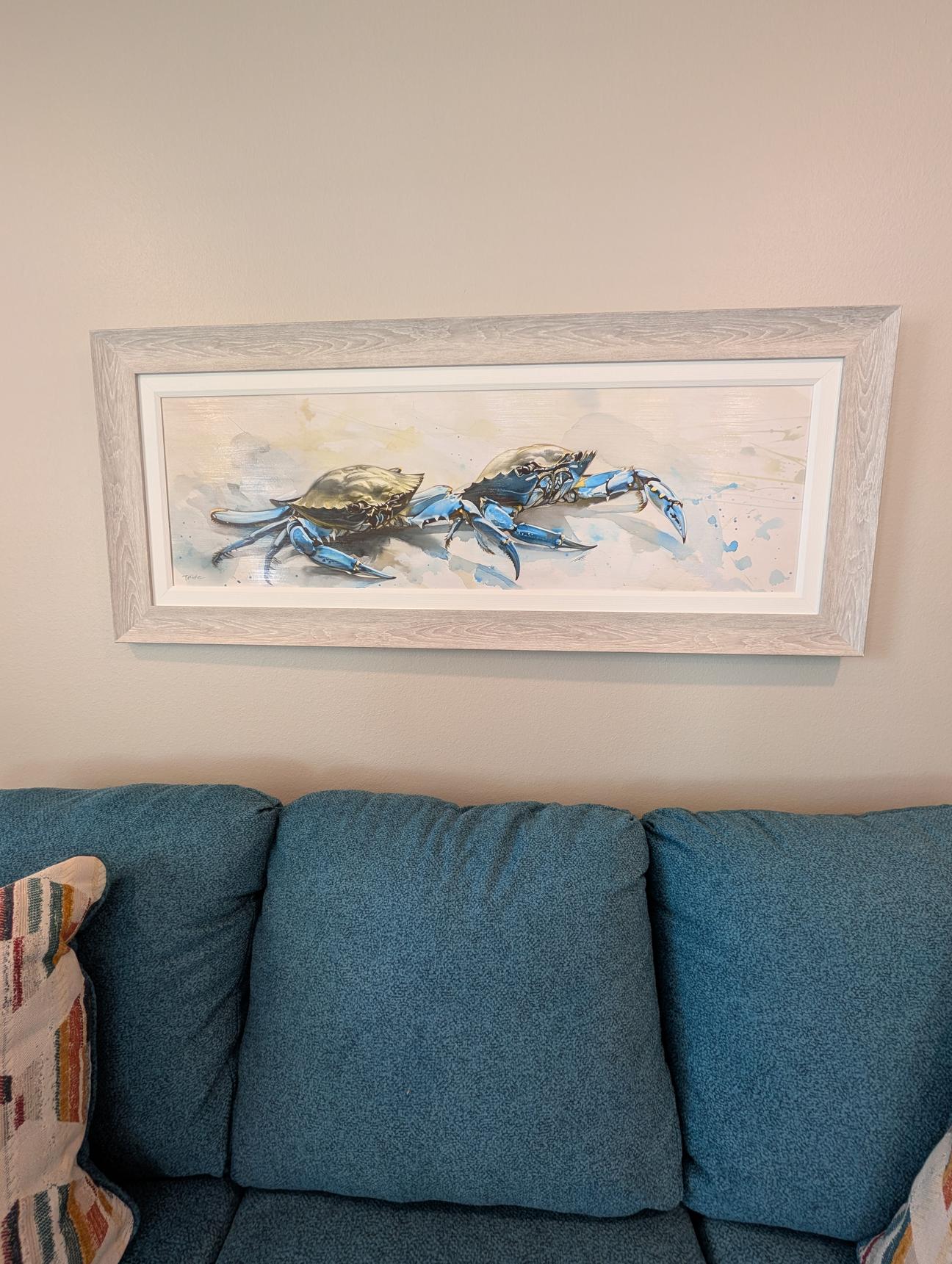 Framed artwork of blue crabs hanging above a blue sofa with decorative pillows against a neutral wall.