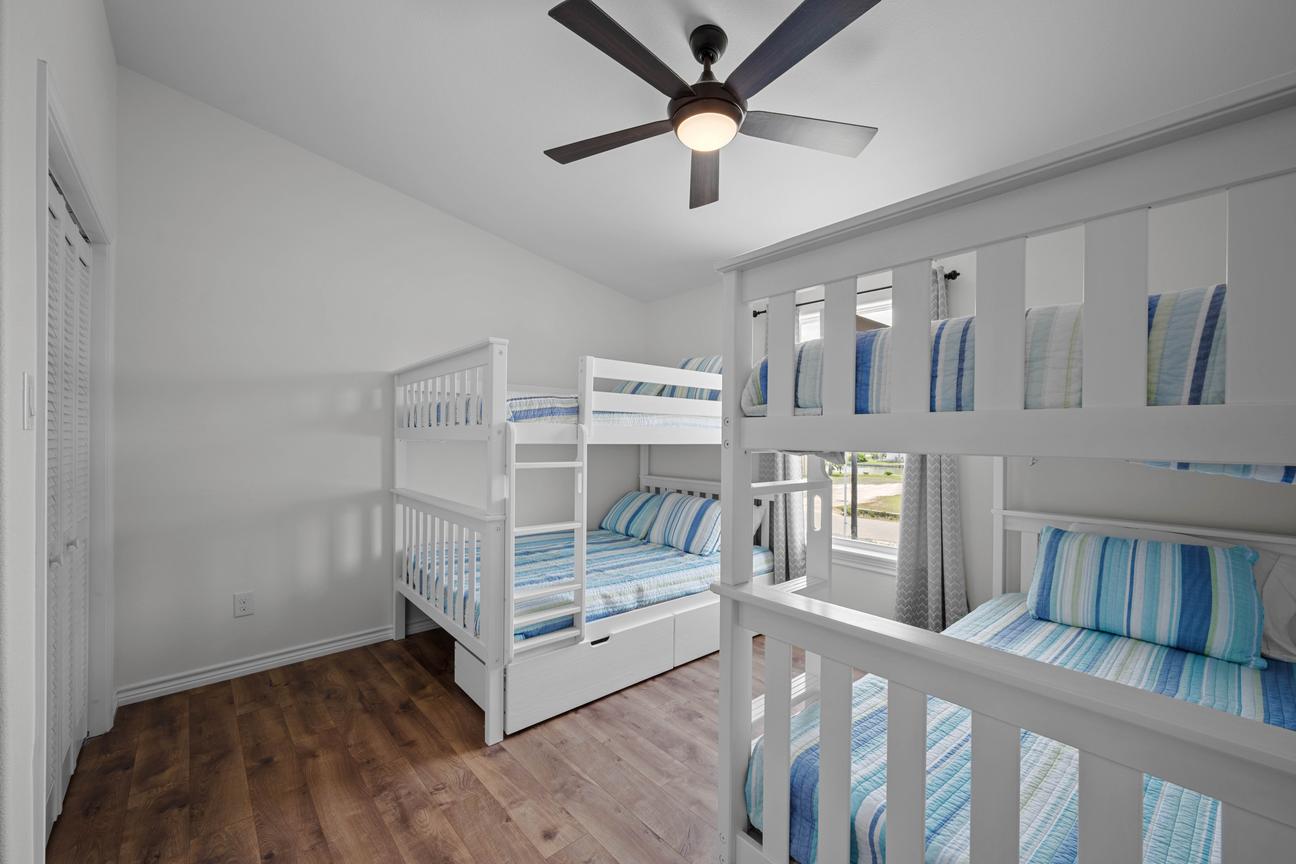Bright bunk room with two white bunk beds, blue striped bedding, wood-look floors, and a ceiling fan.
