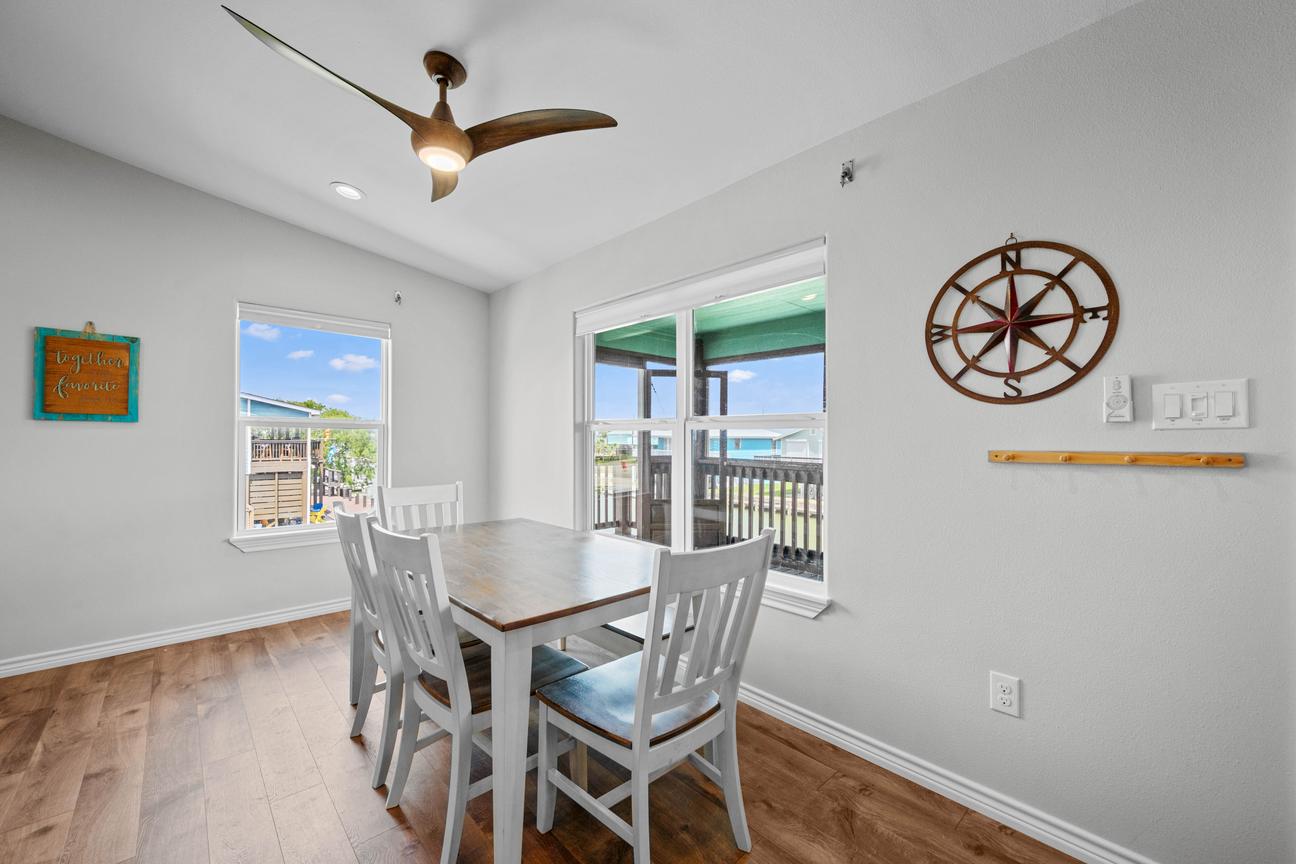 Bright dining area with a wood table, white chairs, nautical wall décor, and glass doors opening to a covered deck.