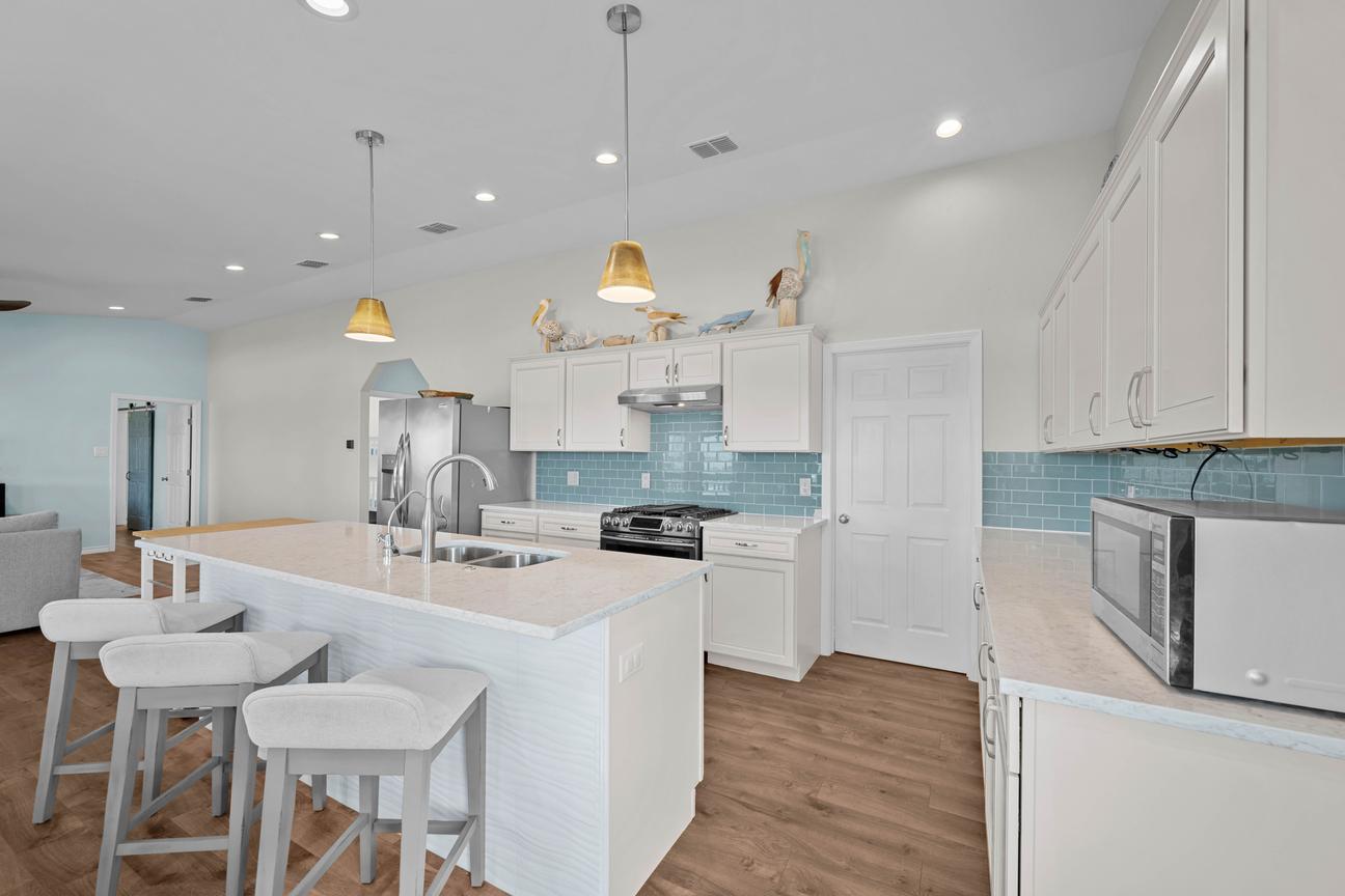Open coastal kitchen with white cabinets, a blue tile backsplash, large island, double sink, and three cushioned barstools.