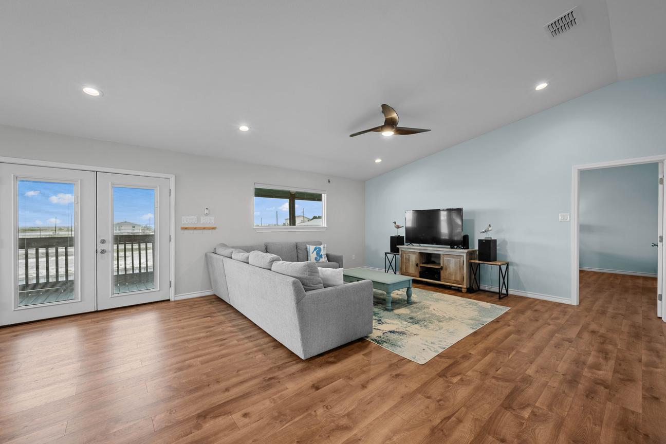 Spacious living room with a gray sectional sofa, large television, coastal blue walls, and glass doors opening to the deck.