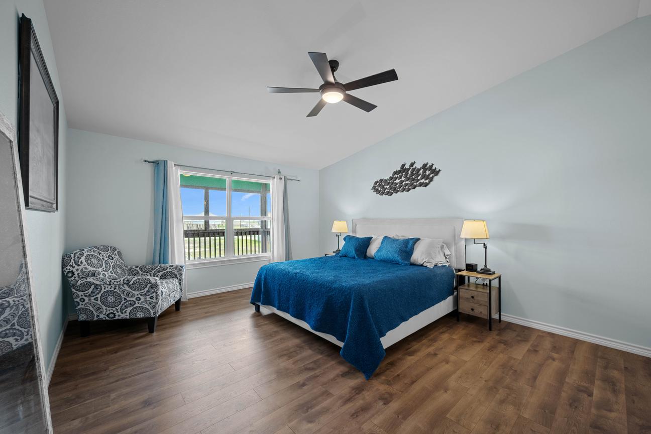 Spacious primary bedroom with a king bed, blue coastal décor, an upholstered chair, wood-look floors, and a ceiling fan.