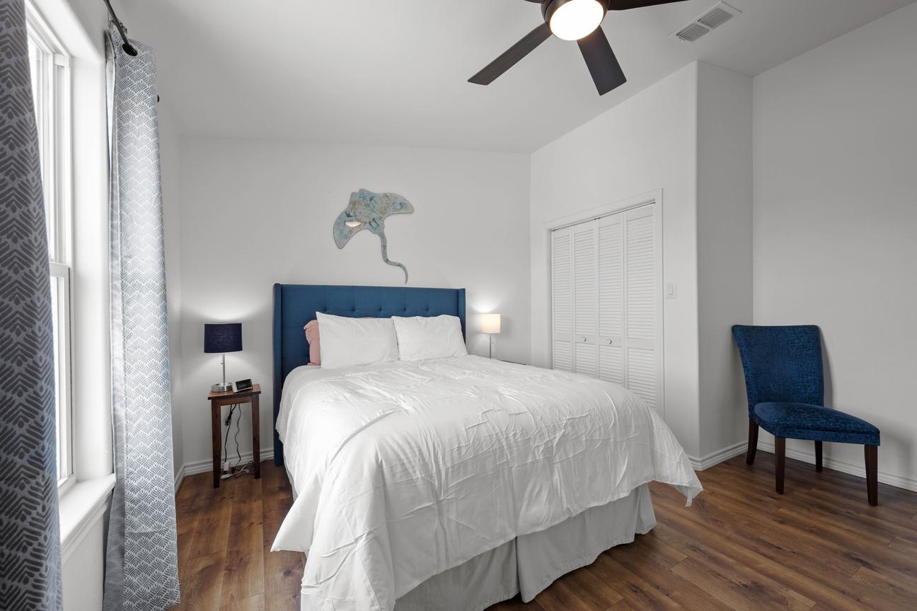 Bright second bedroom with a queen bed, blue upholstered headboard, coastal wall décor, accent chair, and ceiling fan.