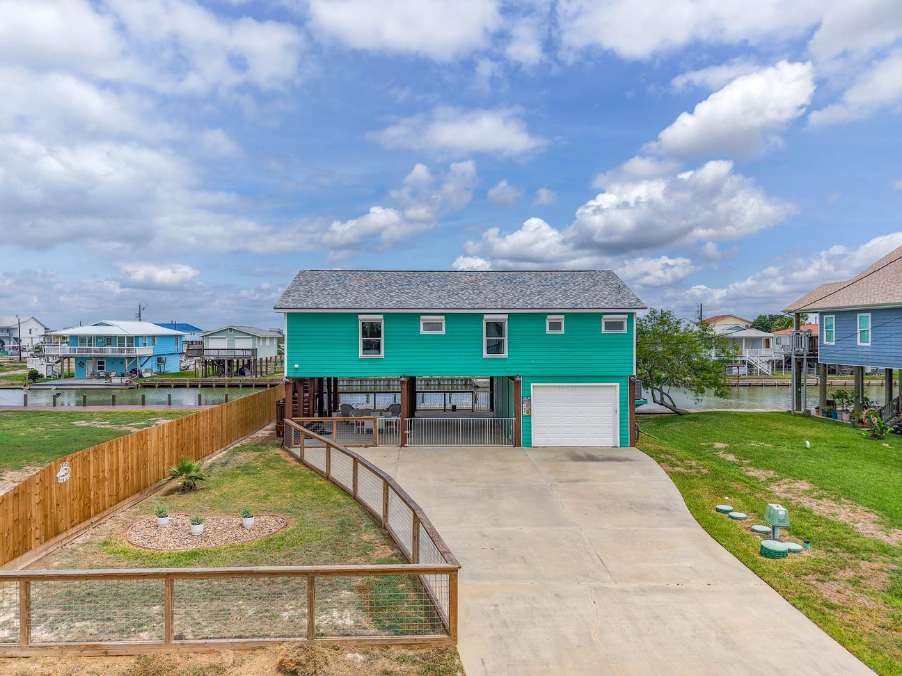 Turquoise raised coastal home with a wide concrete driveway, garage, fenced yard, and covered ground-level patio.