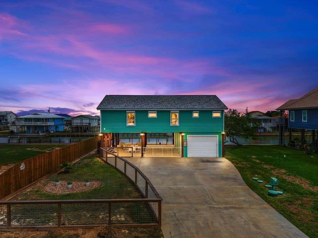 Turquoise raised vacation home glowing at dusk beneath a vivid pink, purple, and blue coastal sky.
