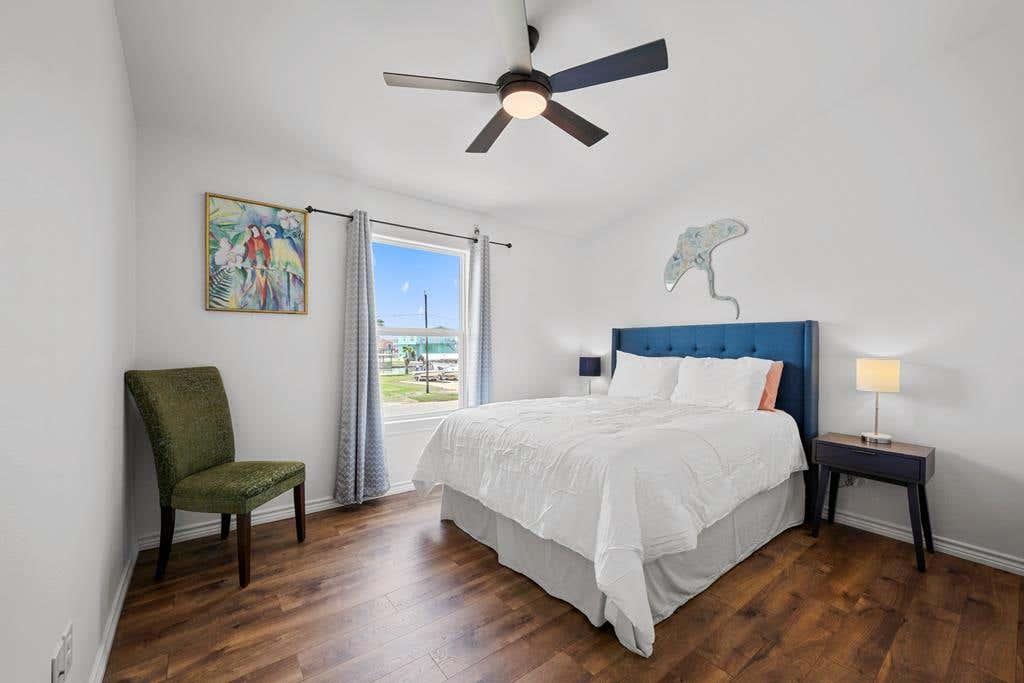 Bright guest bedroom with a queen bed, blue upholstered headboard, coastal artwork, accent chair, and ceiling fan.