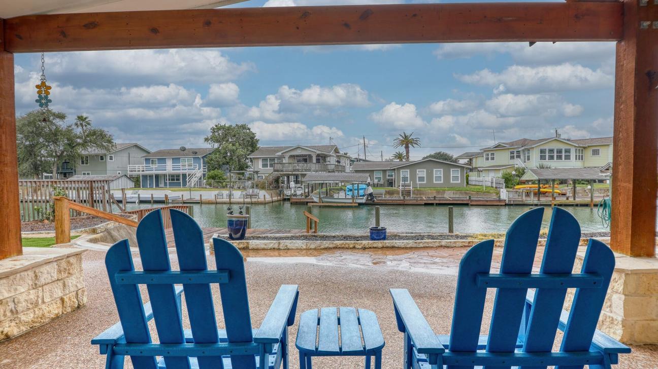 Two blue Adirondack chairs and a small table under a covered patio facing a canal with homes and docks across the water.