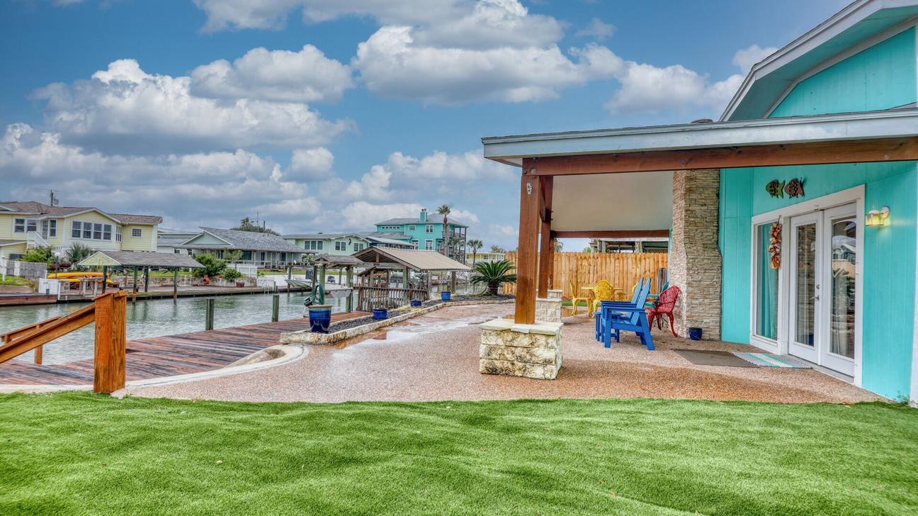 Side view of a turquoise house with covered patio, artificial lawn, and a walkway leading to the canal.