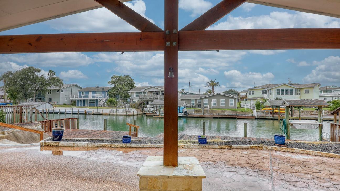 View from under a timber-framed patio cover across a paved patio to the canal and homes on the opposite side.