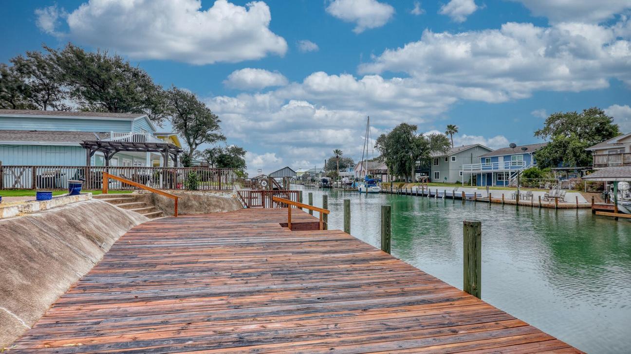 Wooden dock with railings and steps running along a canal, with houses and trees on both banks.