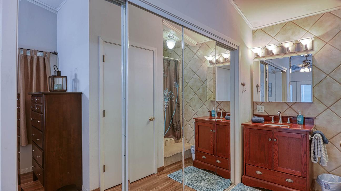 Wooden double vanity area with lighted mirror, mirrored sliding closet doors, and a tall wooden dresser.