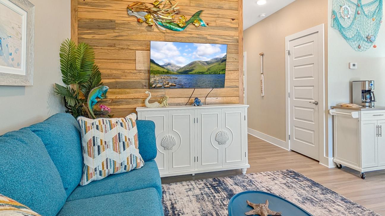 Living room with a blue sofa, wood accent wall, TV on a white cabinet, coastal decorations, and a hallway leading to other rooms.