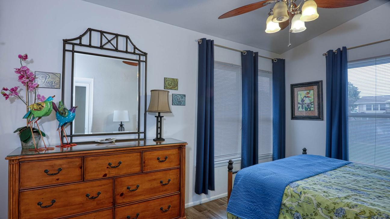 Bedroom with queen bed, blue and green bedding, blue curtains, wooden dresser with mirror, and ceiling fan.