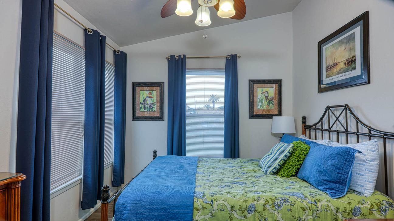 Bedroom with queen bed, blue pillows, black metal headboard, framed art, and windows with blue curtains.