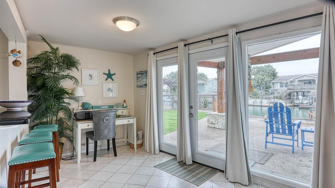 Teal bar stools, a white writing desk with a gray chair, and glass doors opening to a patio with a blue Adirondack chair.