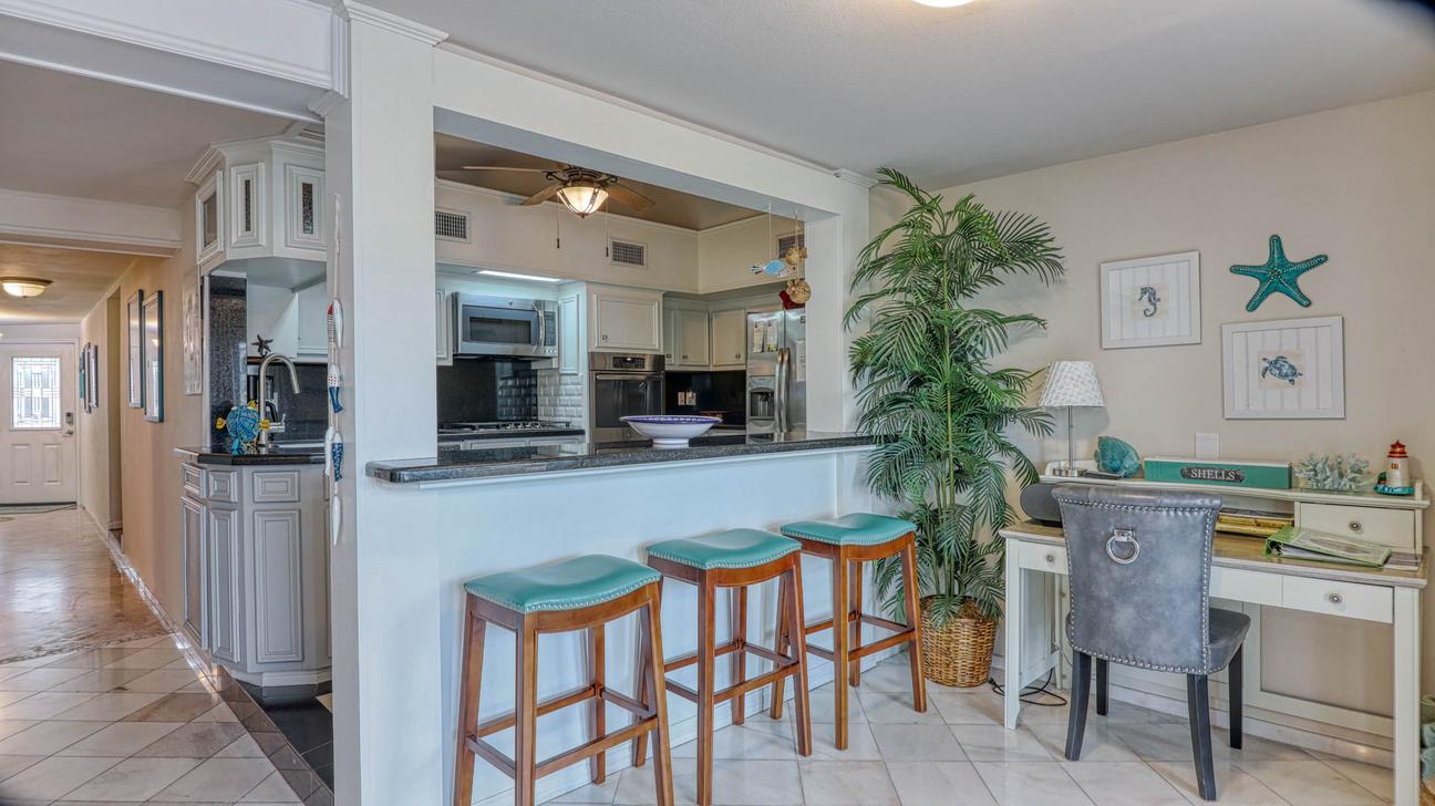 Kitchen with stainless steel appliances, granite breakfast bar with three teal bar stools, and a small white desk with a gray chair.