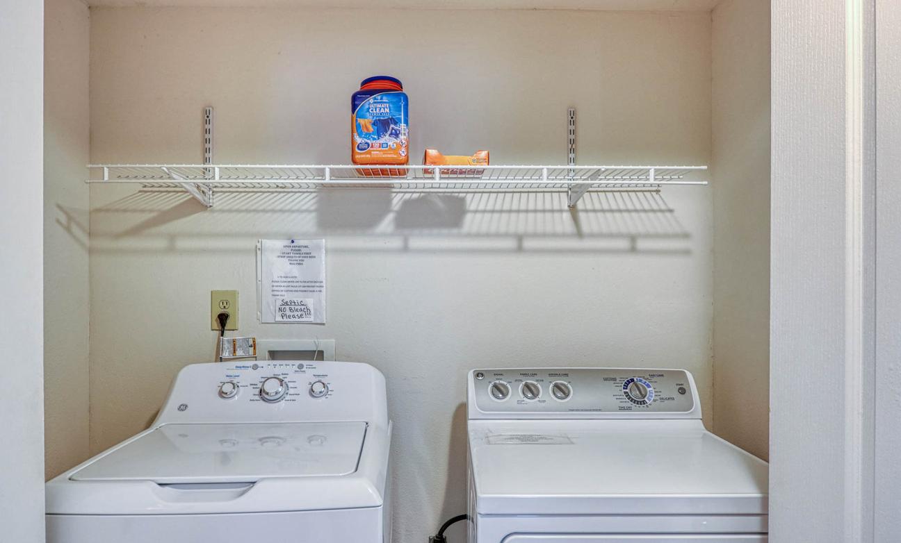 Laundry nook with a white top-loading washer and dryer and a wire shelf above.