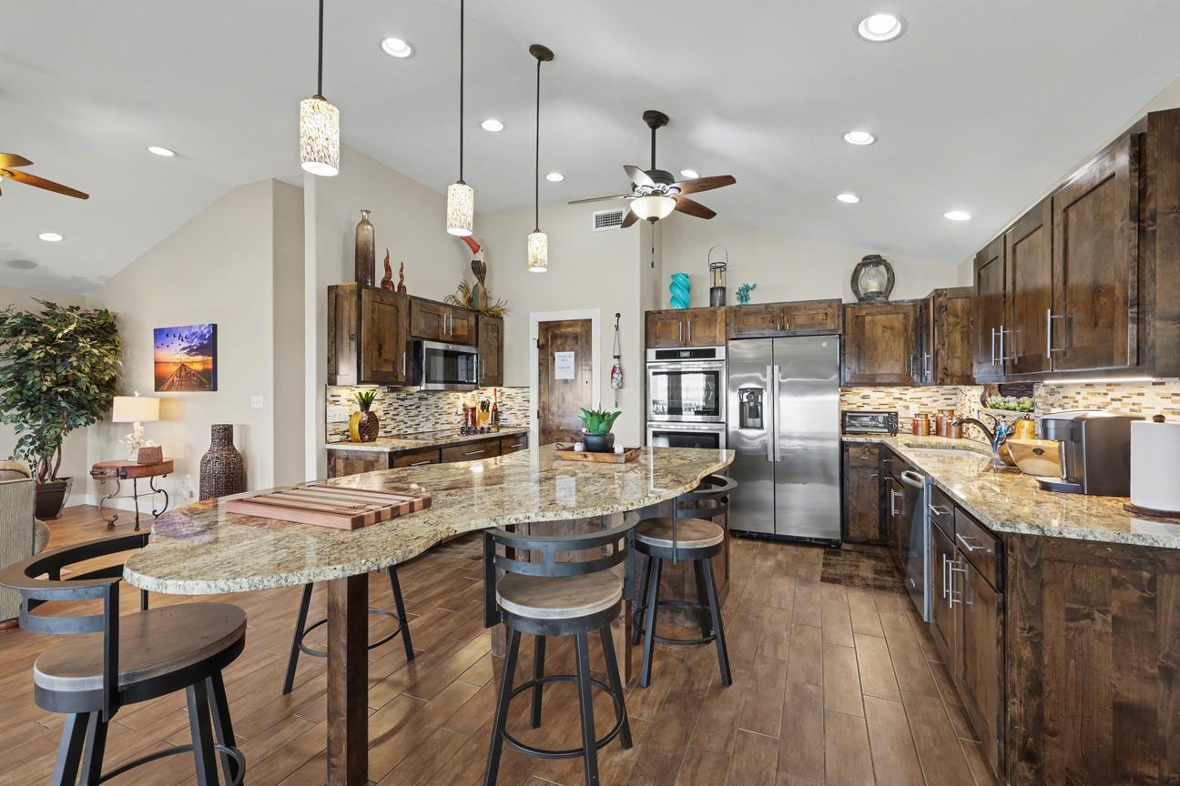 Kitchen with curved granite island and four bar stools, dark wood cabinets, stainless refrigerator, double wall oven, microwave, and pendant lights.