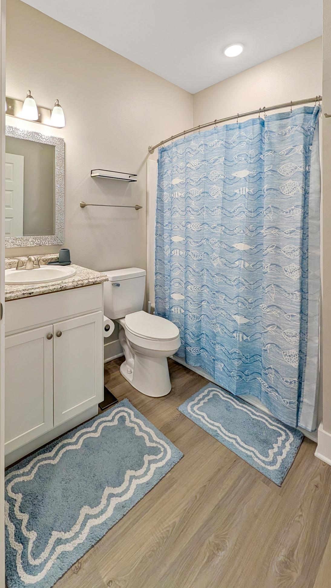 the guest bathrrom has a tub and shower combo with blue coastal shower curtain, blue rug on the floor , white vanity and sink, large mirror above the sink