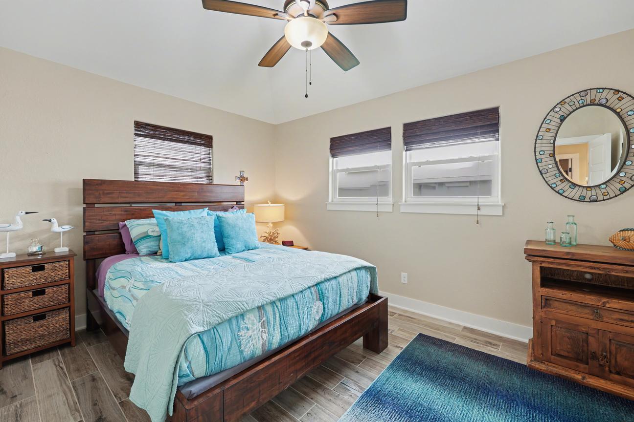 Bedroom with queen bed in a dark wood frame, turquoise bedding, wicker-drawer nightstand, wood dresser, round mosaic mirror, and blue rug.