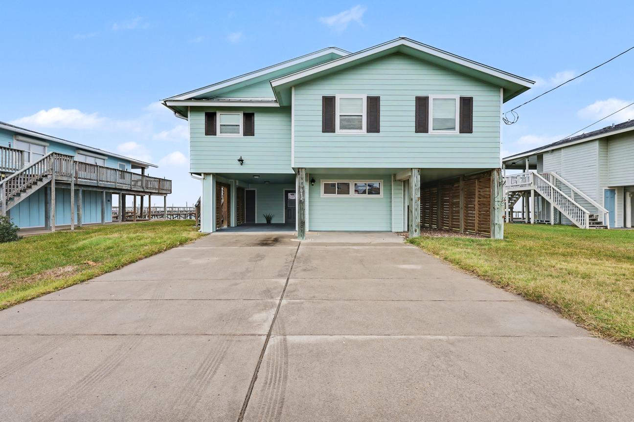 Front of an elevated light green house on pilings, with a concrete driveway, parking area, and lawn on both sides.