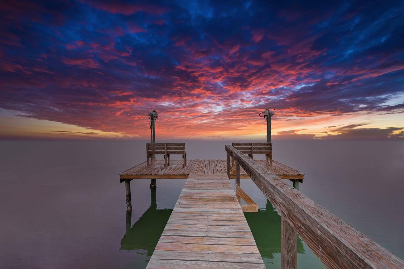 Wooden pier leading to an end platform with two benches and two light posts, under a vivid red and blue sky over still water.