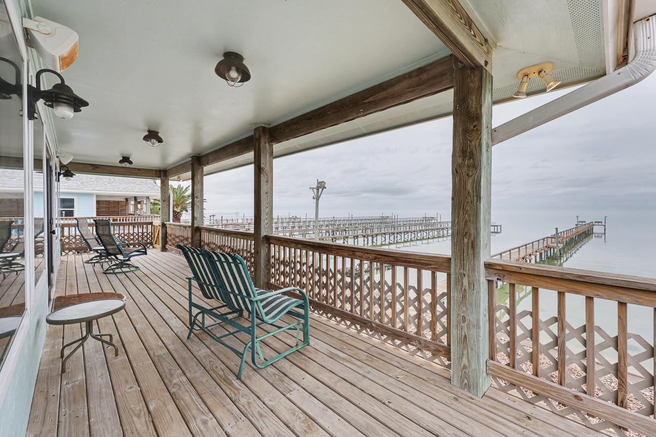 Covered wooden deck with two gliders, lounge chairs, and a small table; lattice railing overlooks bay water and several fishing piers.