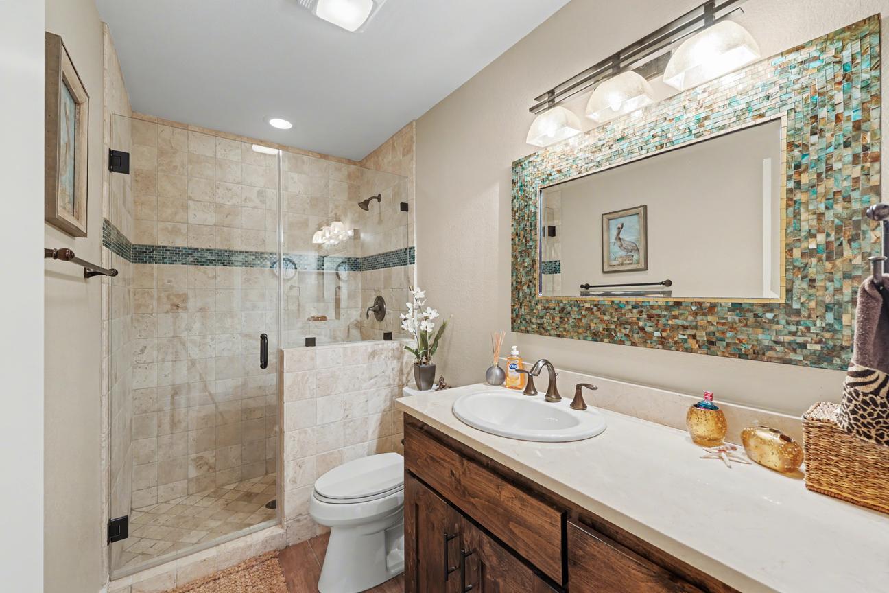 Bathroom with tiled walk-in shower behind a glass door, single-sink wood vanity, toilet, and large mirror framed in blue and gold mosaic tile.