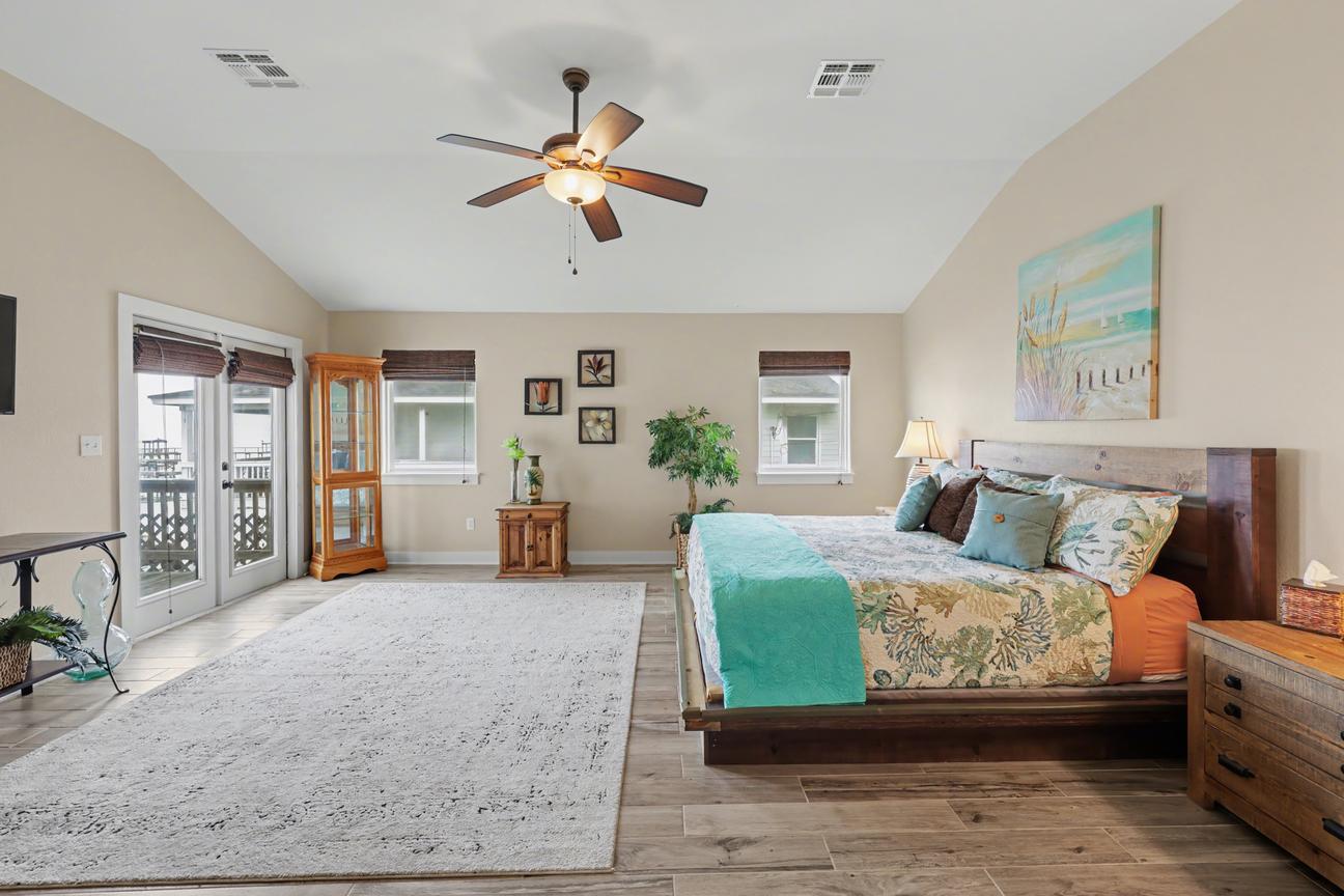 Primary bedroom angle showing king bed, large area rug, curio cabinet, and glass French doors leading to a deck with railing.