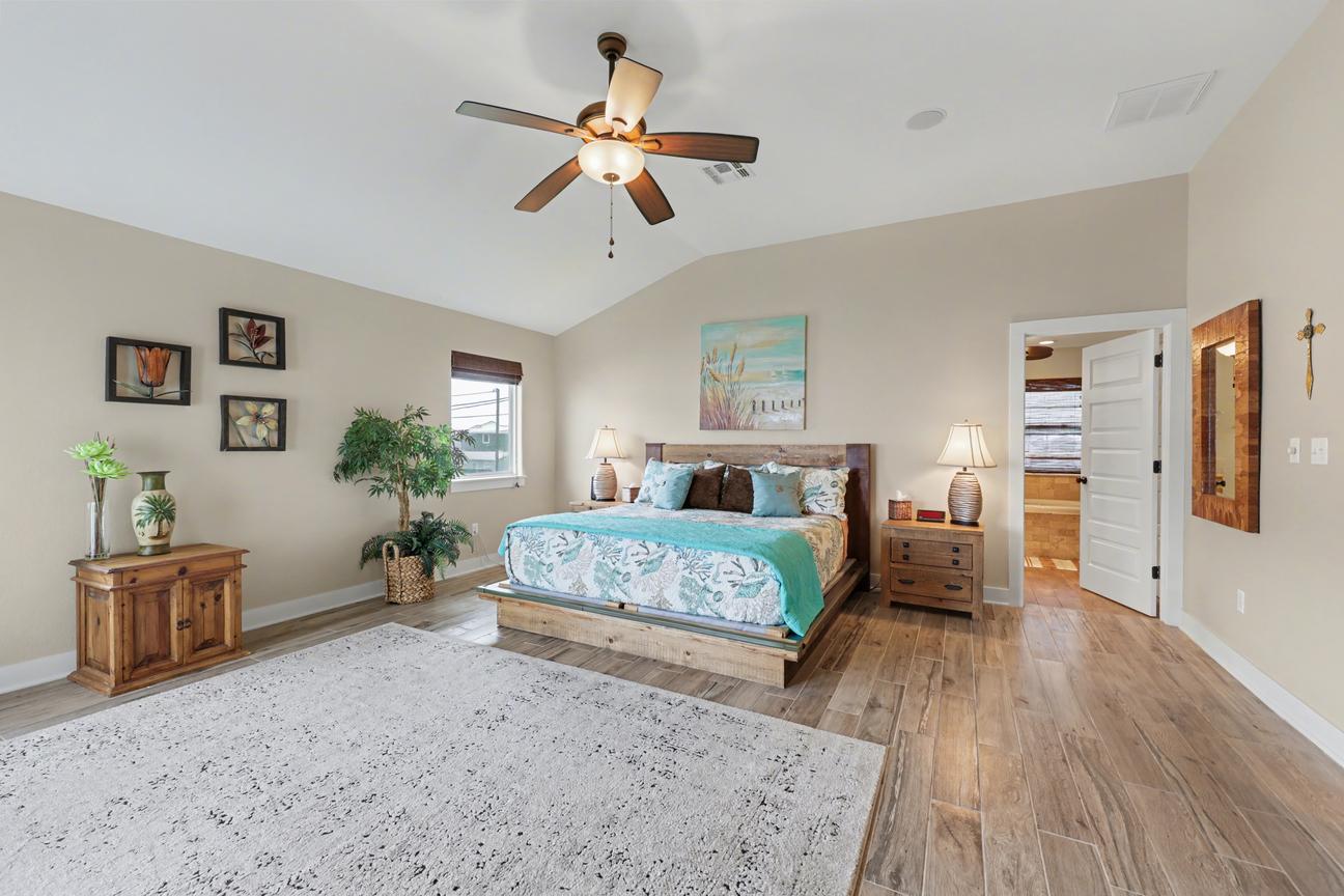 Primary bedroom with king platform bed, two nightstands with lamps, vaulted ceiling with fan, area rug, and an open door to a bathroom.
