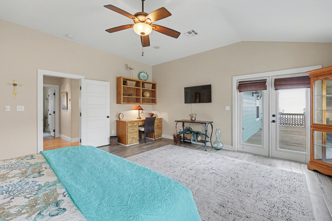 View from the foot of the king bed toward a wood desk, wall-mounted TV, curio cabinet, and French doors opening to a deck.
