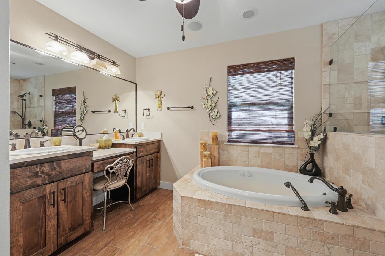 Bathroom with two-sink wood vanity, vanity stool, large mirror, and a tiled drop-in tub beneath a window with a woven shade.