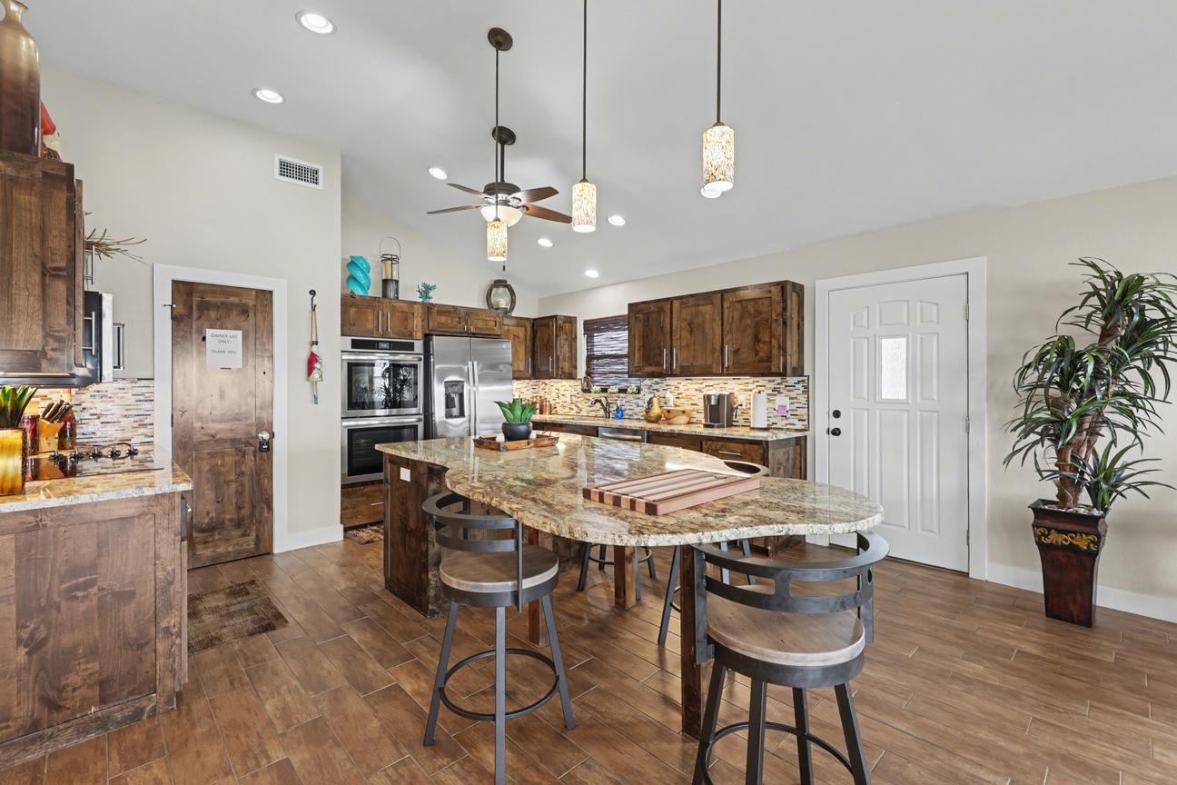 Kitchen side view with granite island and bar stools, stainless double wall oven and refrigerator, a wood interior door, and a white exterior door.