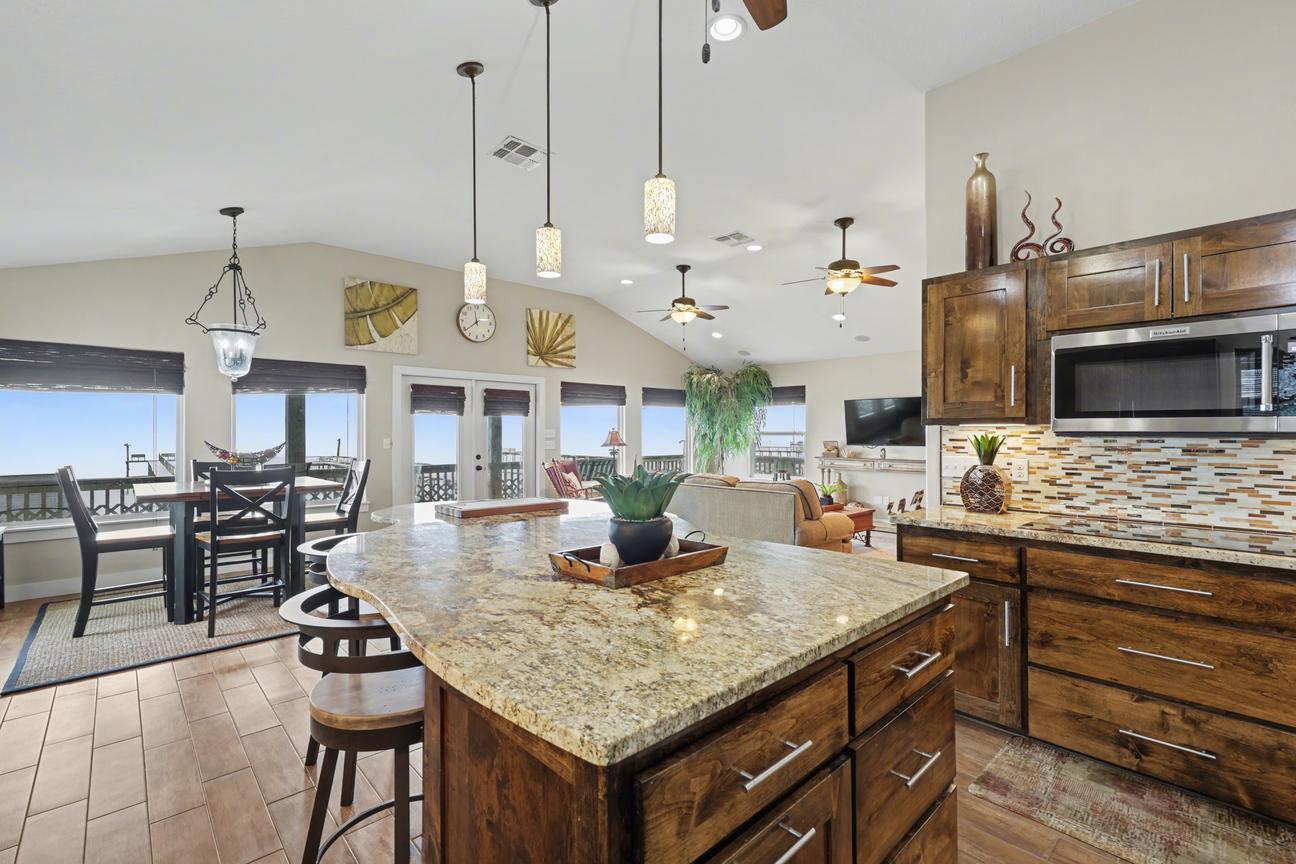Granite kitchen island with bar stools in the foreground, looking past the dining table to French doors, windows, and the living area.