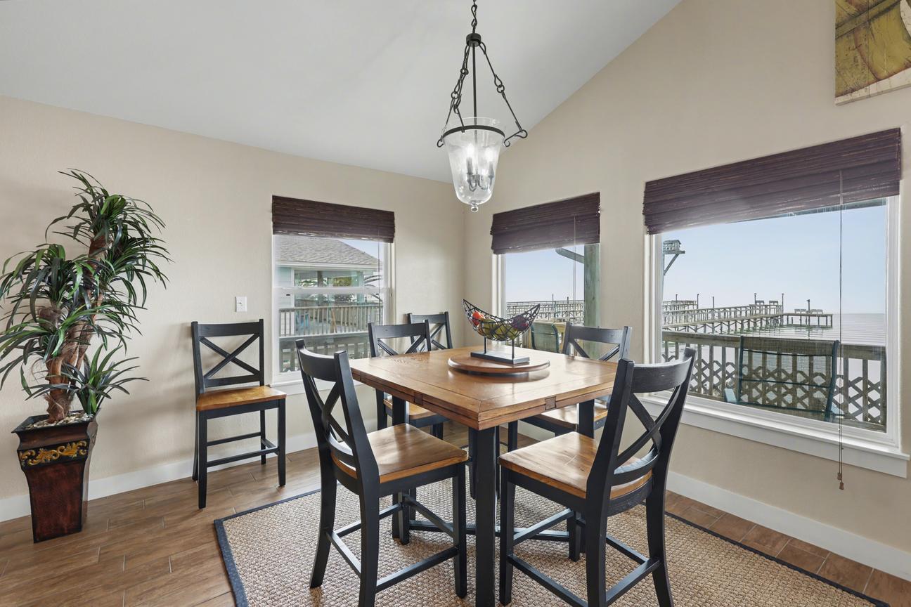 Dining area with high-top wood table and six black chairs, pendant light, and windows showing a deck railing, piers, and water.