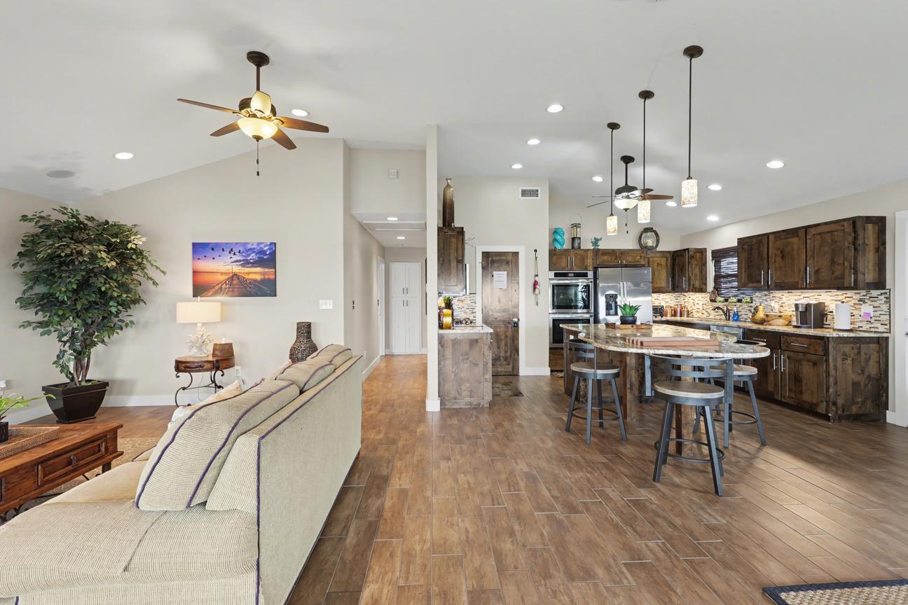 Open floor plan view from behind the living room sofa toward the kitchen island, bar stools, stainless appliances, and a hallway.
