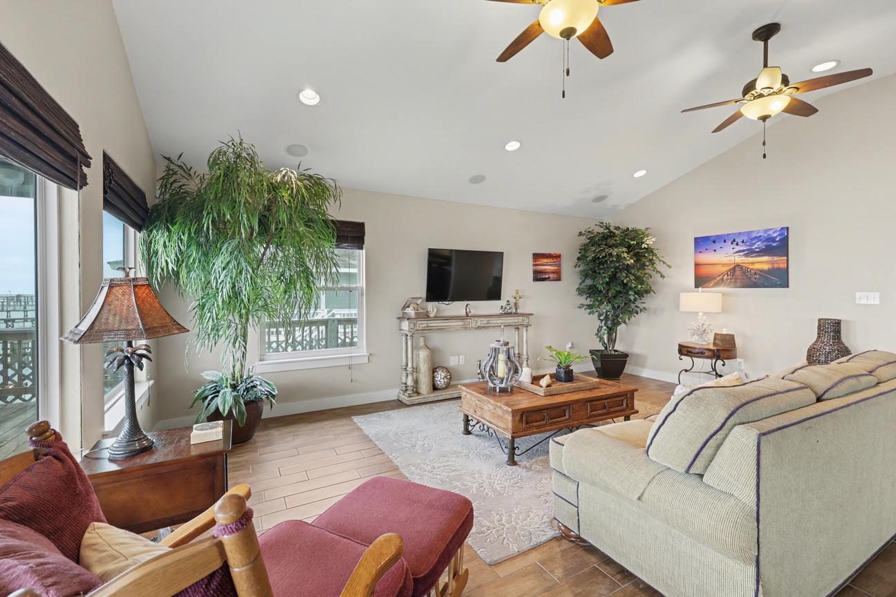 Living room angle showing wall-mounted TV over a console table, sofa, rocking chair with ottoman, coffee table, and large indoor plants.