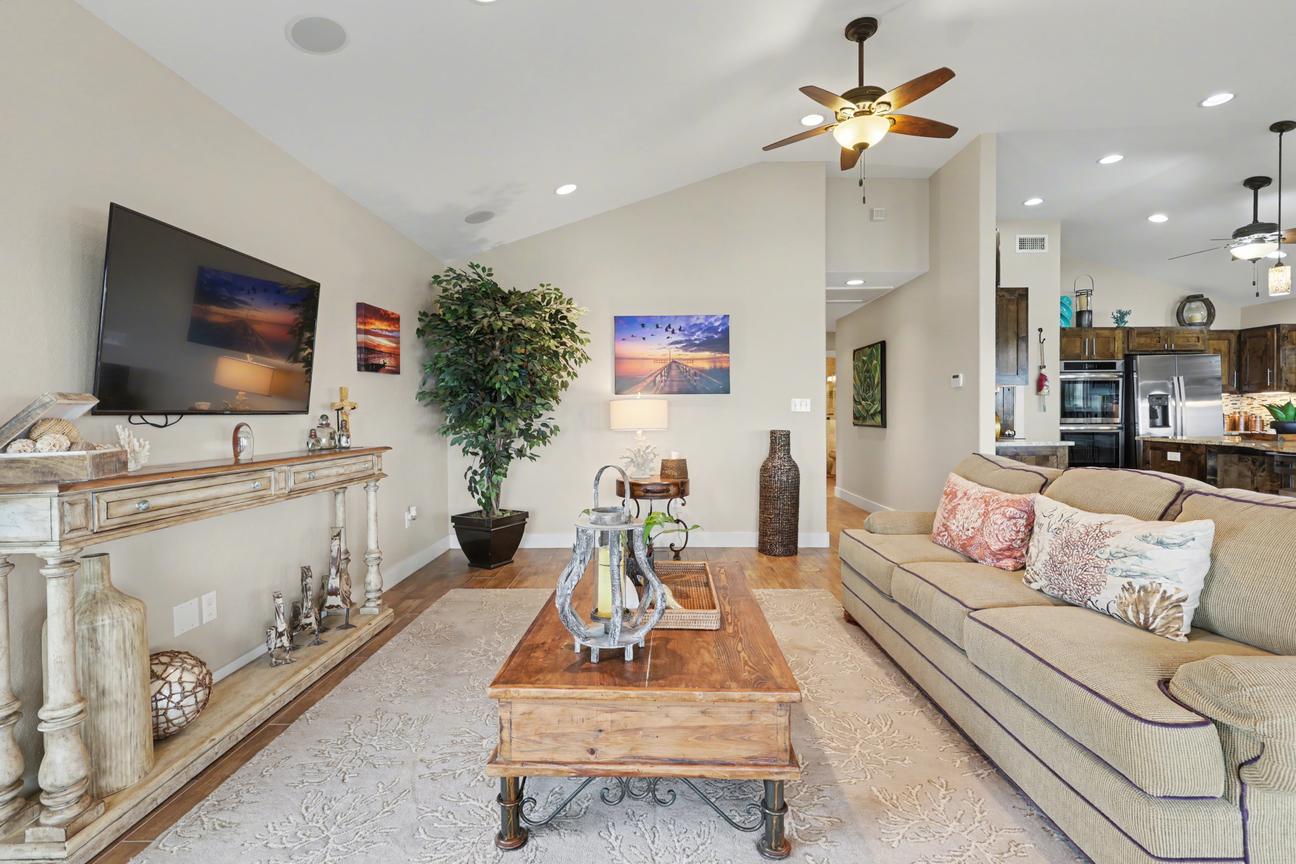 Living room with wall-mounted TV above a carved console, sofa with patterned pillows, wood coffee table, and the kitchen visible beyond.