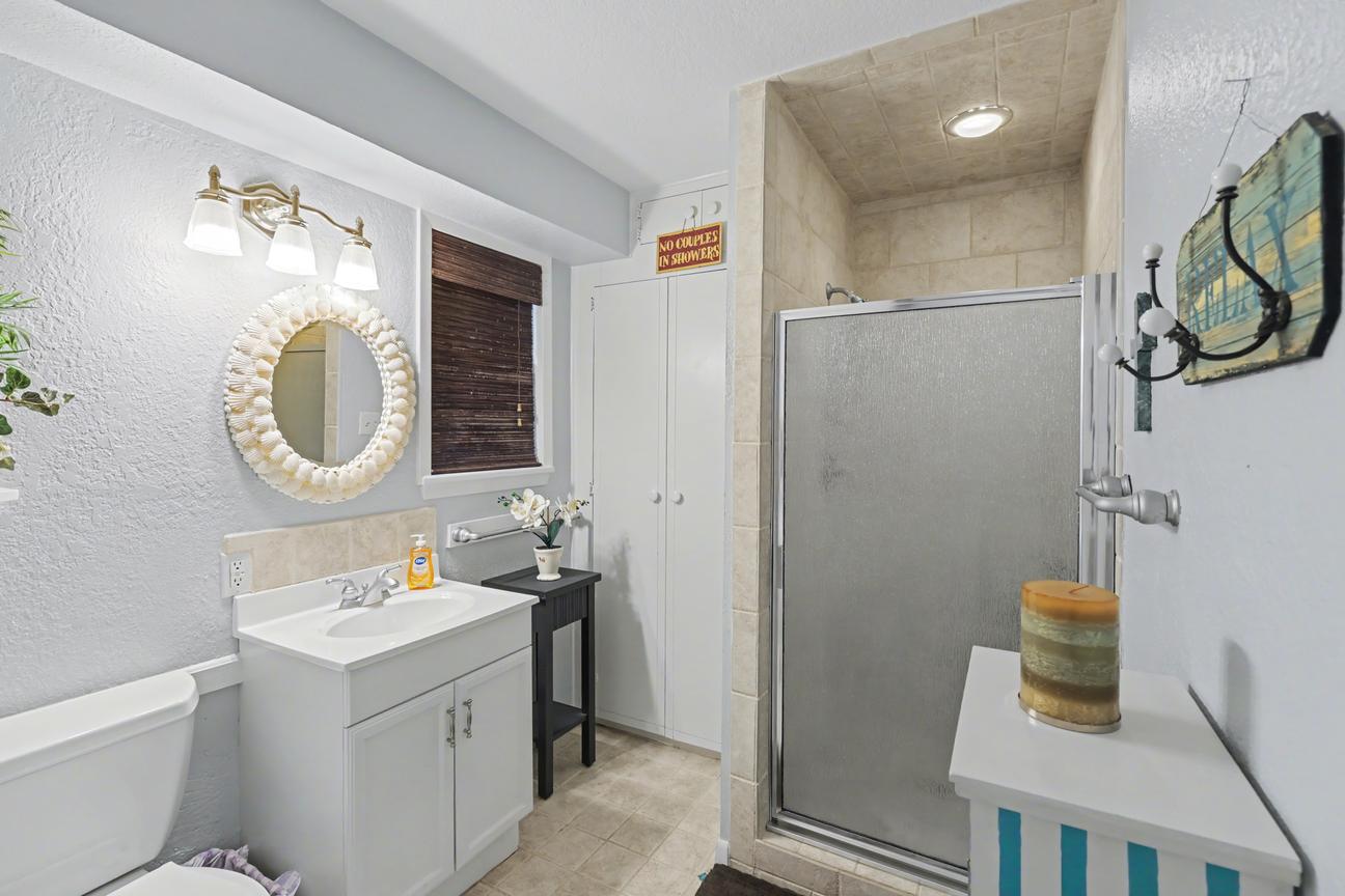 Bathroom with white single-sink vanity, shell-framed round mirror, glass-door shower stall, white cabinet, and toilet.