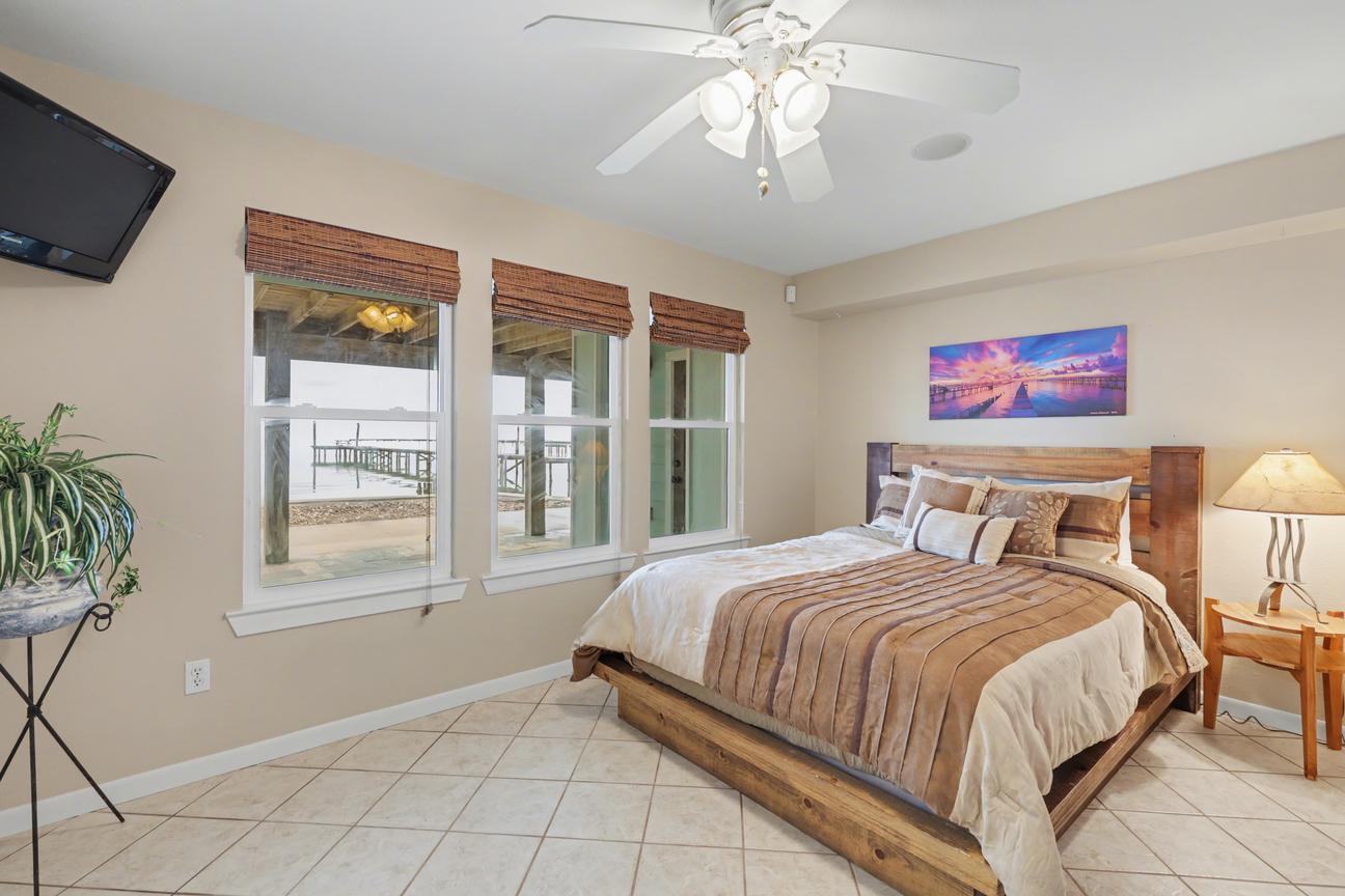 Bedroom with queen bed in a wood frame, wall-mounted TV, ceiling fan, tile floor, and three windows showing piers and bay water.