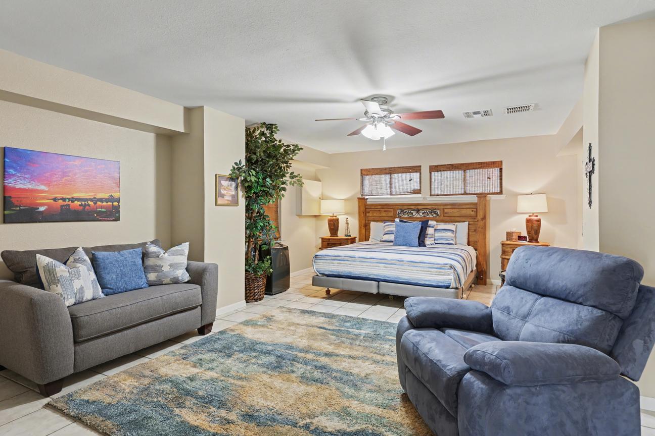 Large bedroom with tile floor, bed with wood headboard and nightstands, gray loveseat, blue recliner, area rug, and tall indoor plant.