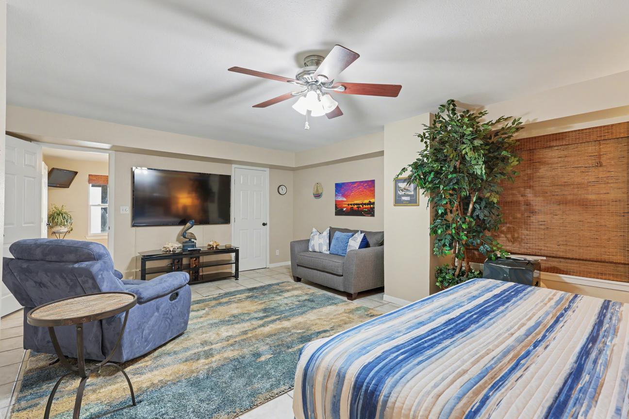 Bedroom sitting area with large wall-mounted TV on a media stand, gray loveseat, blue recliner, side table, and doorway to another room.