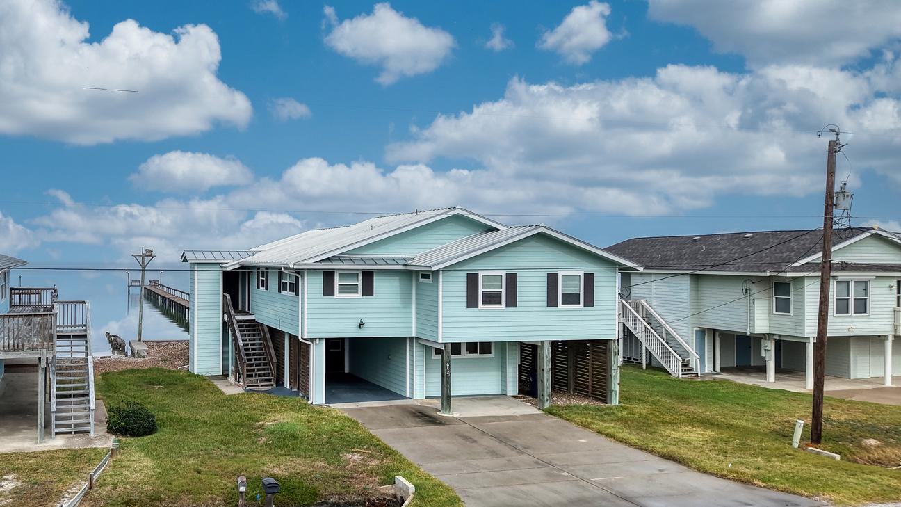 Elevated light green house with metal roof, exterior stairs, covered parking, and driveway; bay water and a pier are visible behind it.