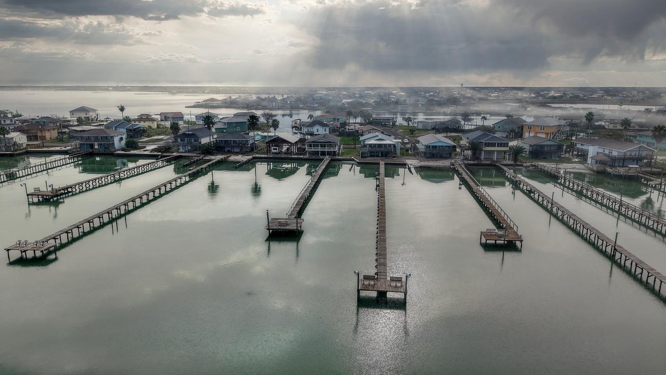 Aerial view of several long wooden piers extending into calm bay water from a row of waterfront homes under cloudy skies.