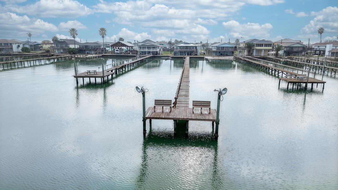 Aerial view of a pier end T-head with two benches and two light fixtures over green bay water, with houses and palm trees on shore.