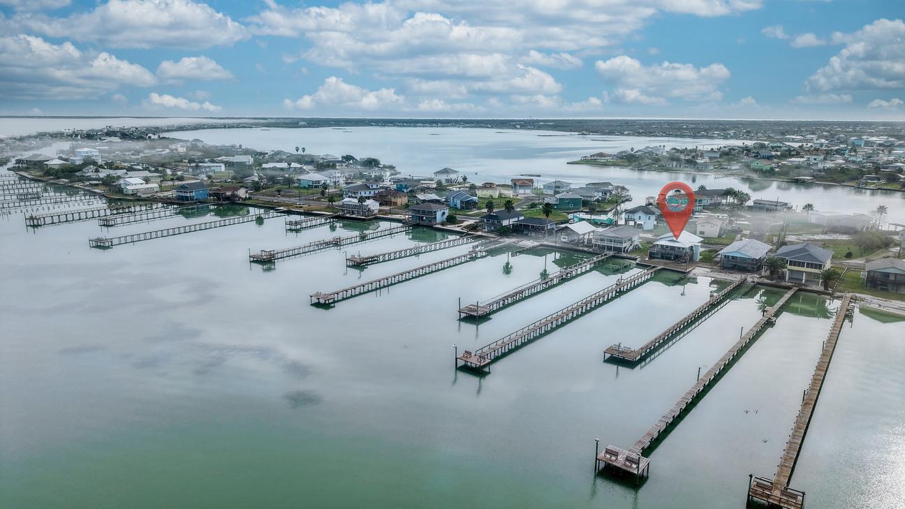 Aerial view with a red location pin over a waterfront house, with many piers extending into the bay and shoreline homes beyond.