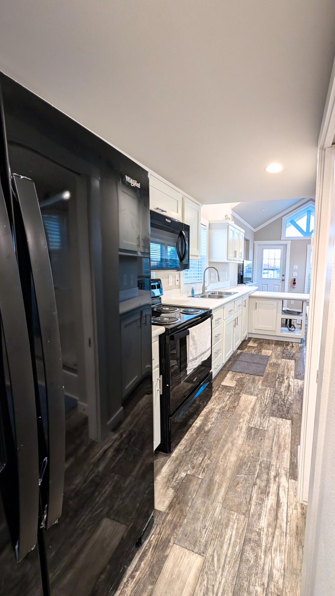 Galley-style kitchen with black refrigerator and stove, white cabinetry, wood-look flooring, and a view toward the open living area with natural light.