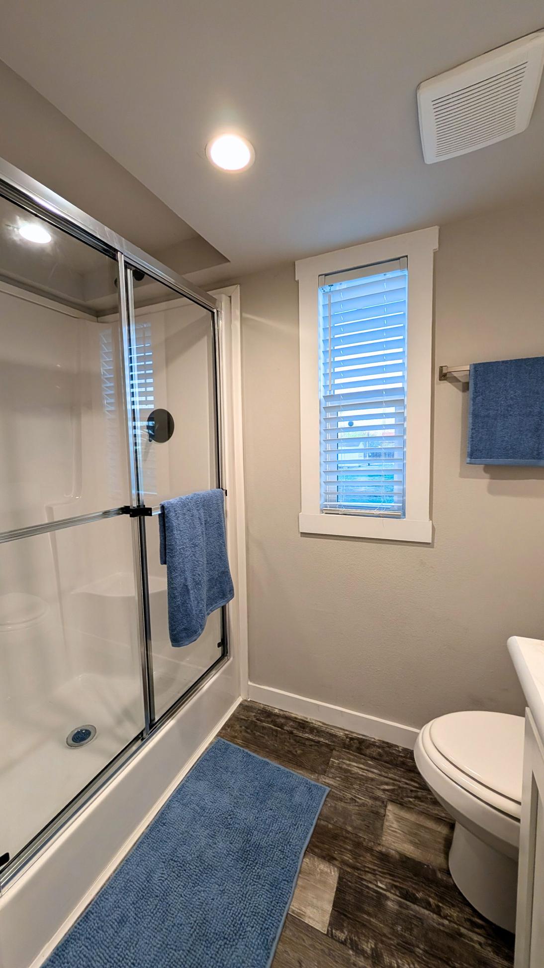 Bathroom with a glass-enclosed walk-in shower, white walls, window with blinds, blue towels, and wood-look flooring.