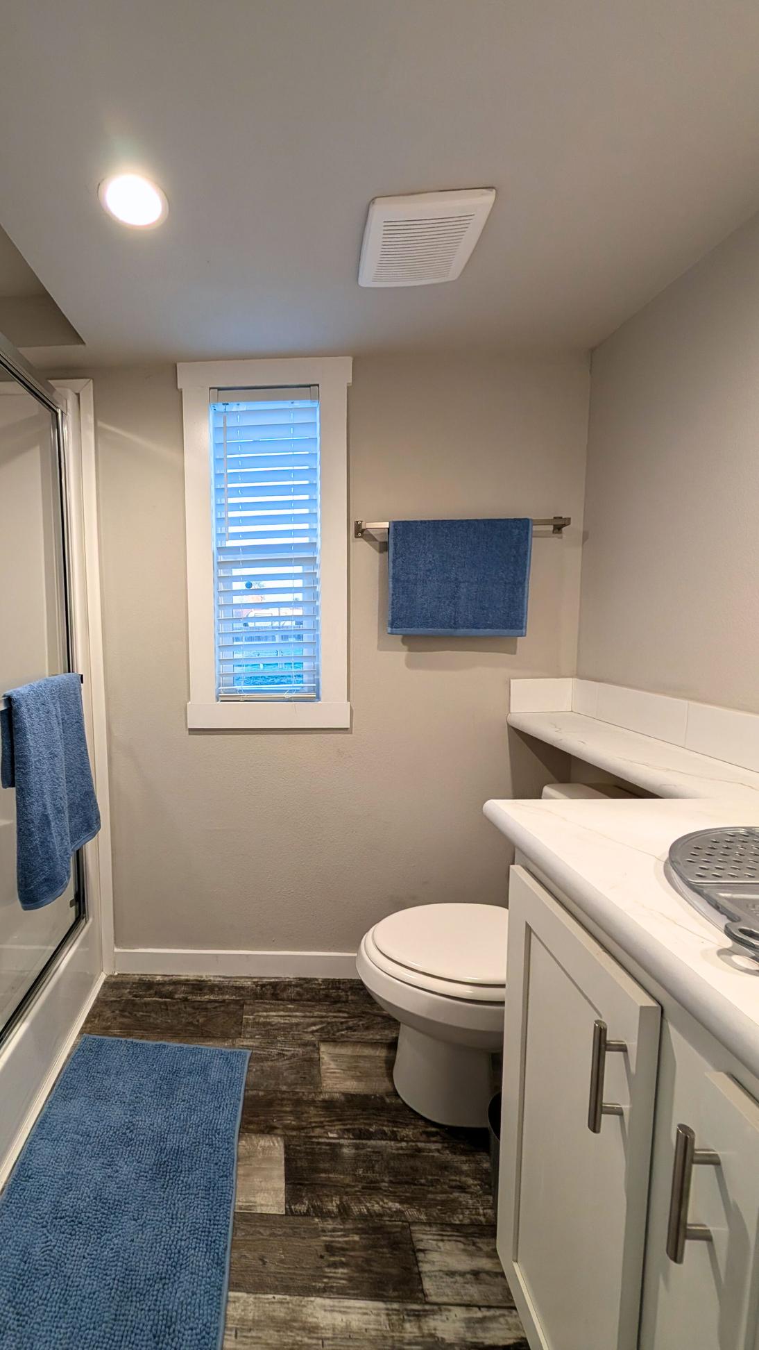 Bathroom with white vanity and countertop, toilet, window with blinds, blue towels, and a glass-enclosed shower on the side.